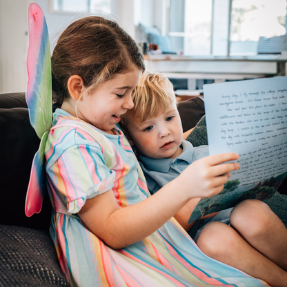 Two children sitting on a couch and reading a fairy letter. The older child wears colourful fairy wings, and the younger child wears a light blue shirt.